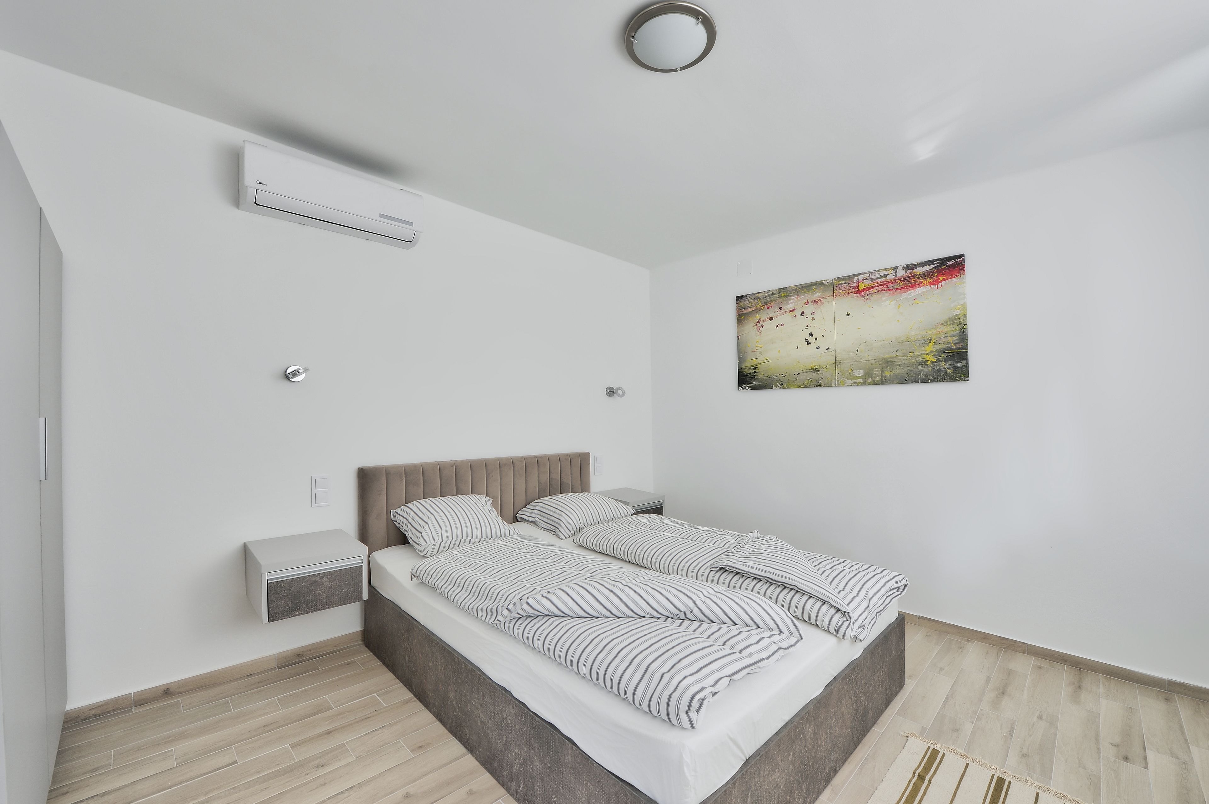 Un dormitorio moderno con cama, mesita de noche, equipo de aire acondicionado y un cuadro en la pared.