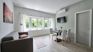 A modern living room with a sofa, dining table, and chairs. There are large windows with curtains, a flat-screen TV mounted on the wall, and a door. - Villa Oliver 2 Siófok (Siófok)