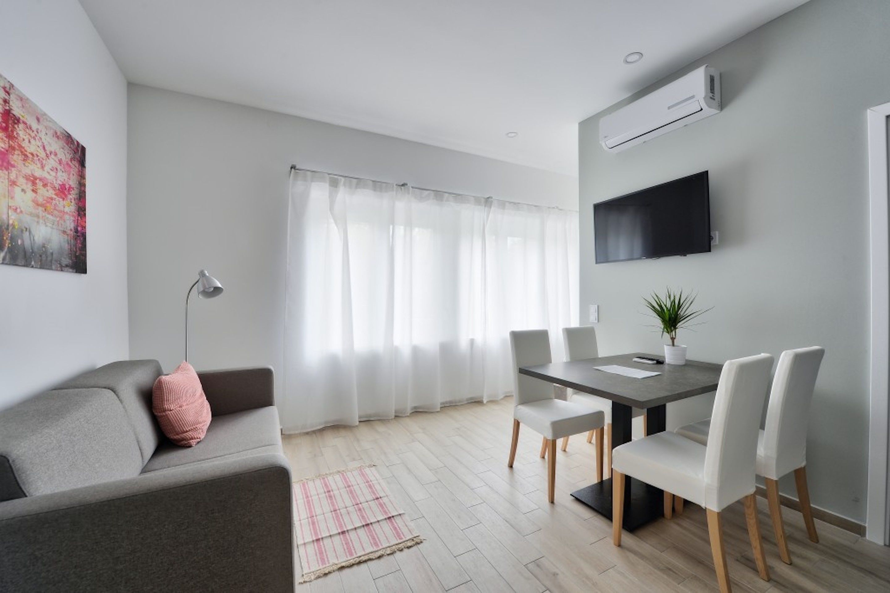 Un salon moderne avec un canapé gris, une table à manger entourée de chaises blanches, un téléviseur à écran plat fixé au mur et une grande fenêtre ornée de rideaux légers.