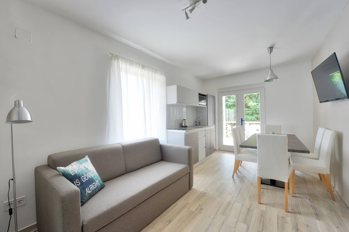 A modern living room with a sofa, a dining table with chairs, a kitchen area, and a TV mounted on the wall.