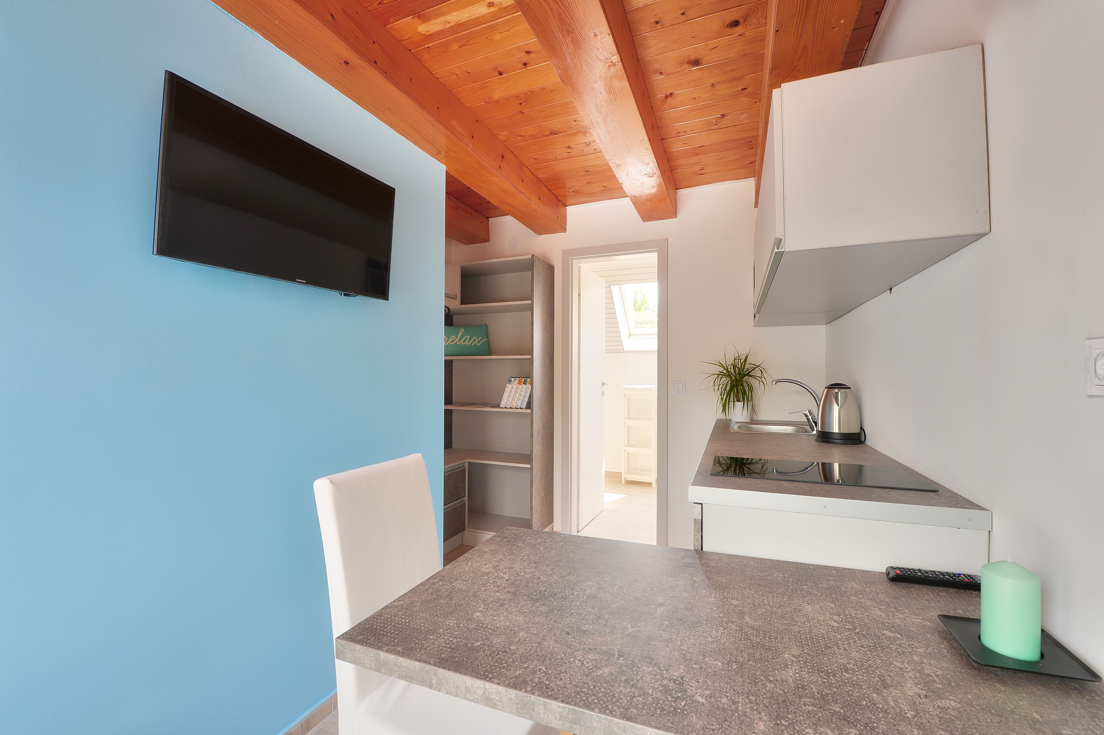 Cocina compacta con estufa integrada, pileta y una pequeña mesada. Hay una silla y un televisor montado en la pared.