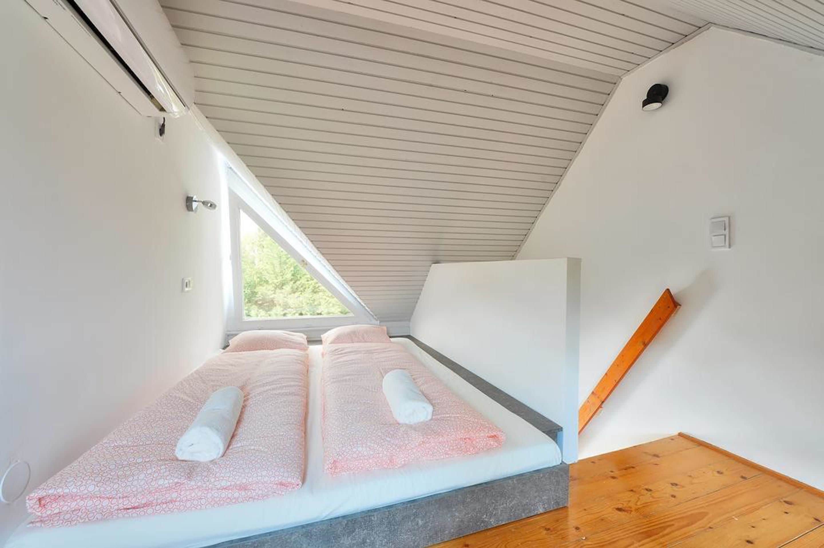 Una habitación con techo inclinado, una cama con ropa de cama rosa y un ventanal que deja entrar luz natural.