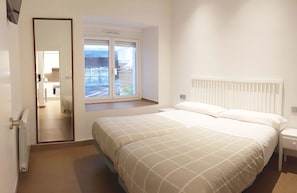 Quarto com cama, mesa de cabeceira, janela com persianas e porta para um banheiro.