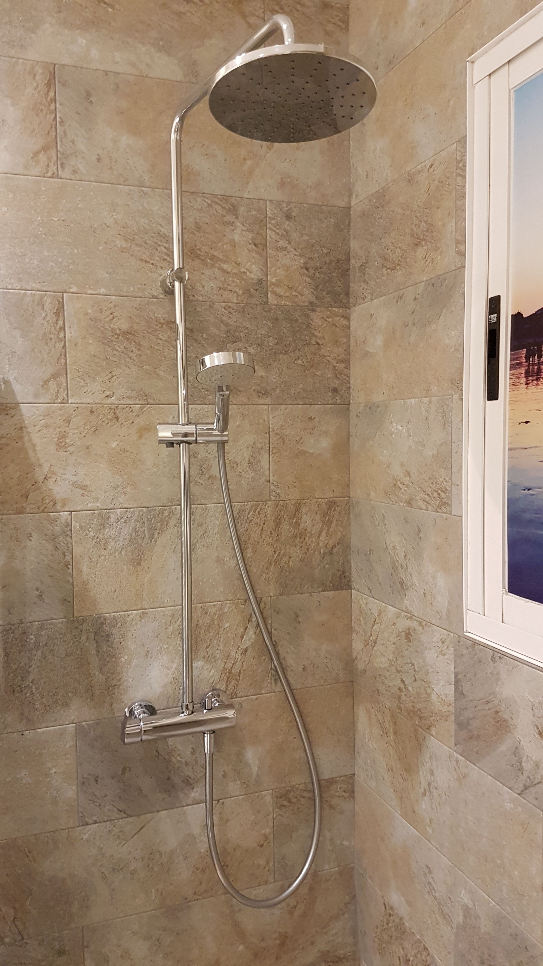 A shower with a handheld showerhead and a fixed rain showerhead, mounted on a wall with beige tiles.