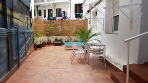 Une cour avec un sol carrelé, une table entourée de chaises, des plantes en pot et un fil à linge suspendu.