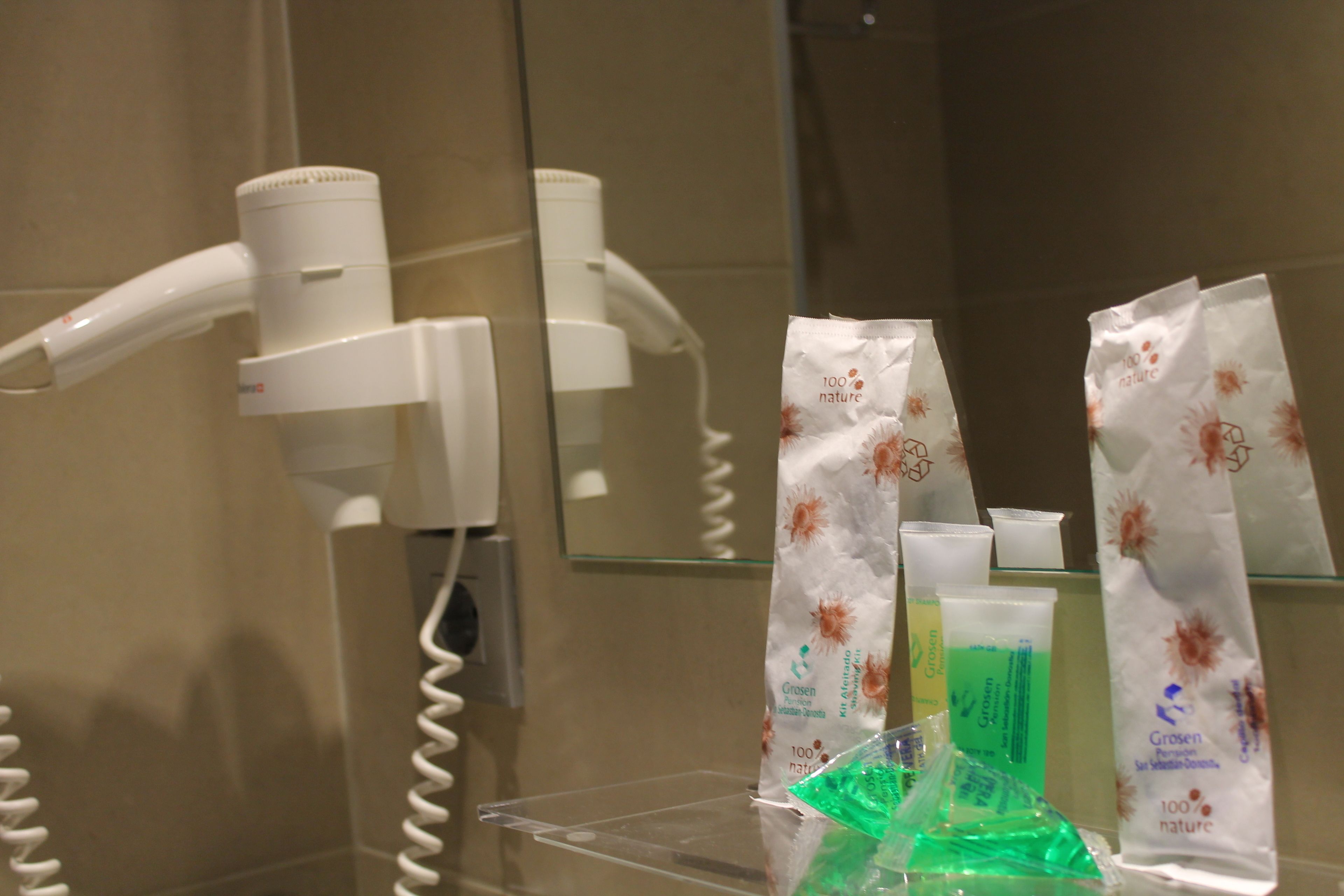 A bathroom with a hairdryer, a mirror, and toiletries including a green bottle and a white bag with orange floral patterns.