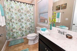 A bathroom with a shower curtain, toilet, sink, and a window with blinds. - 4412AC (Davenport)