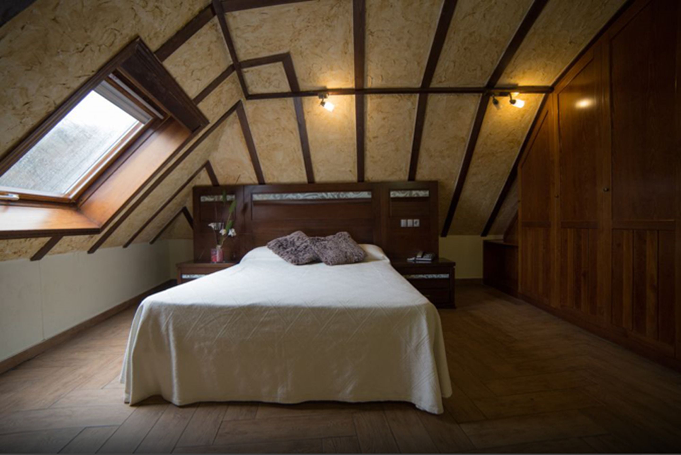 A bedroom with a sloped ceiling, a skylight, a bed, a nightstand, and a wooden wardrobe.