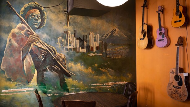 A mural of a person playing a guitar, a round wooden table, and several guitars on the wall.
