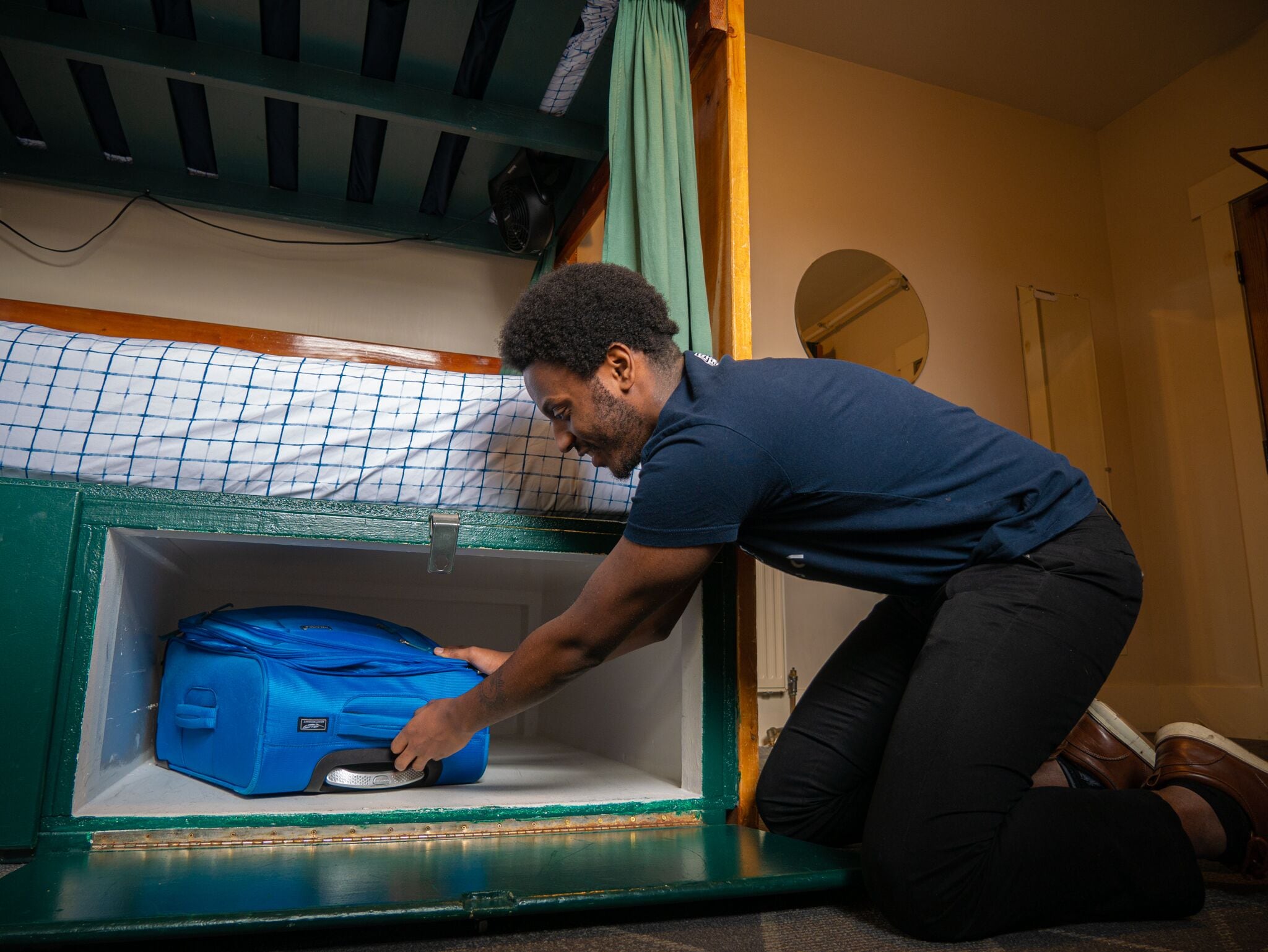 A person placing a blue suitcase into a green storage compartment under a bunk bed.