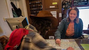 A person wearing a plaid shirt and a cowboy hat sitting at a table with a map, another person in a denim jacket, and a computer monitor in the background.