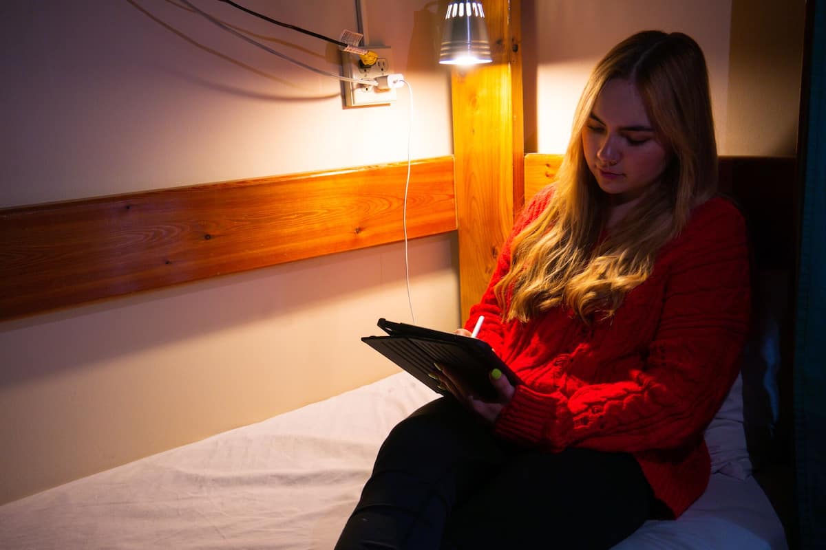 A person sitting on a bed in a room with a wooden headboard, a wall socket, and a lamp.