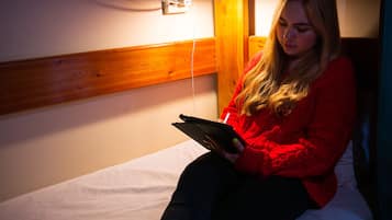 Una persona sentada en una cama en una habitación con cabecera de madera, un enchufe y una lámpara.