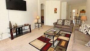 A living room with a flat-screen TV, a glass coffee table, a sofa with patterned cushions, a side table with a lamp, and a rug with geometric patterns. - 1842 NHD (Davenport)