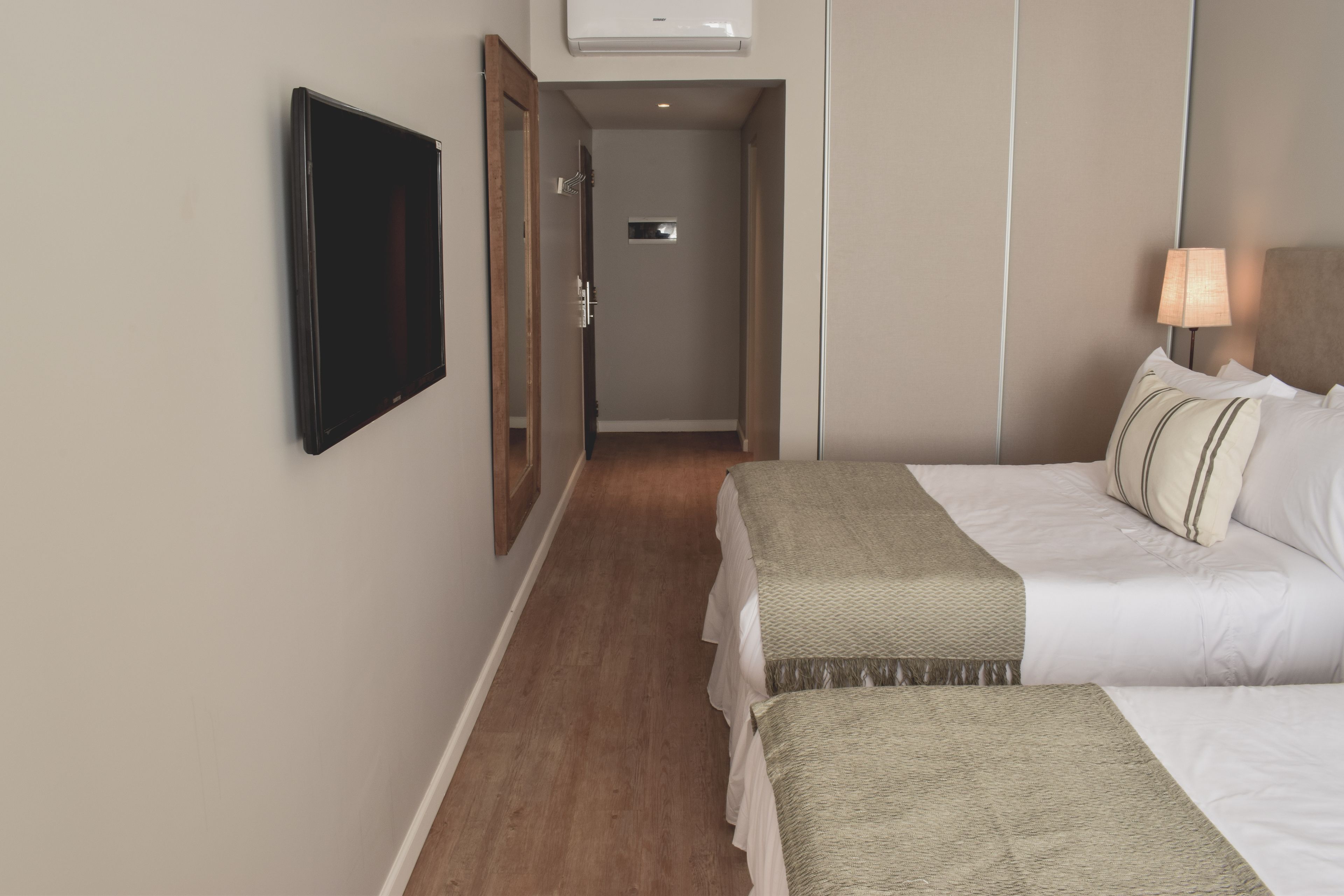 A hotel room with two beds, a flat-screen TV mounted on the wall, a bedside lamp, and a wooden floor.