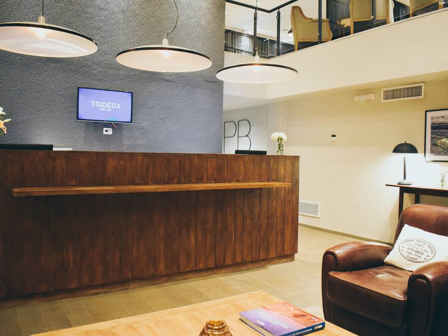 Un mostrador de recepción con una pantalla de televisión mostrando “TRIDEOA”, una barra de recepción de madera, un sillón de cuero marrón con un cojín, y una mesa de centro de madera con un libro y un jarrón de flores.