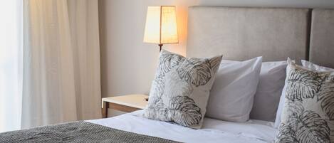 Una cama bien hecha con un edredón gris, dos almohadas con motivos de hojas, una lámpara de noche y un cuadro en la pared.