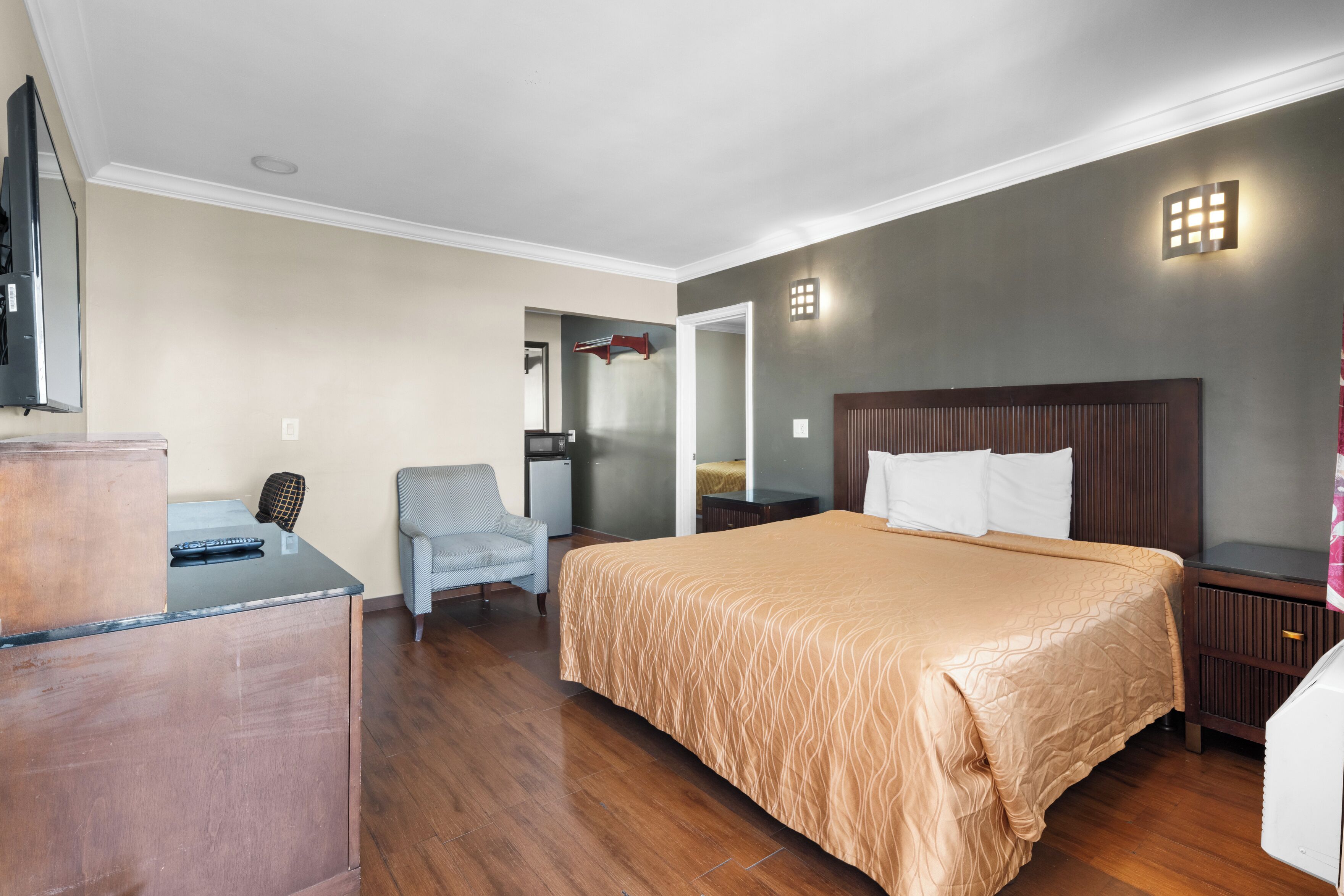 A hotel room with a large bed, a desk, a chair, a TV, and a small kitchenette.