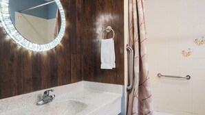 Un baño con tocador de madera, espejo redondo con luces, ducha con cortina y un inodoro blanco.