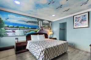 A bedroom with a mural of a coastal scene, a bed with a patterned bedspread, a nightstand, and a framed picture on the wall. - Orange Villa Park Motel (Orange)