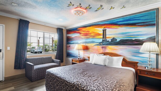 Habitación de hotel con una cama grande, un sofá, una mesita de noche con lámpara y un cuadro de un atardecer sobre un faro.