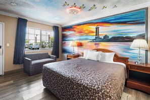 A hotel room with a large bed, a sofa, a nightstand with a lamp, and a painting of a sunset over a lighthouse. - Orange Villa Park Motel (Orange)
