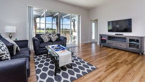 Una sala de estar moderna con un sofá negro, una mesa de centro de vidrio, un televisor en un mueble gris y un amplio ventanal corredero que da a un patio con vistas a palmeras.