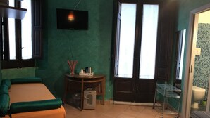 A room with a bed, a small table with a cup and a kettle, a television on the wall, and a door leading to a bathroom. - L'Angolo sulla Piazza (Mirabella Imbaccari)