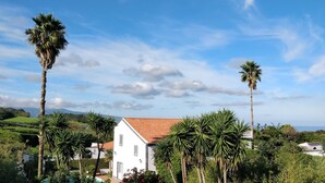 A white building with a red roof, surrounded by palm trees and greenery, with a clear sky and distant hills. - Quinta Minuvida Orchard Lodge (Ribeira Grande)