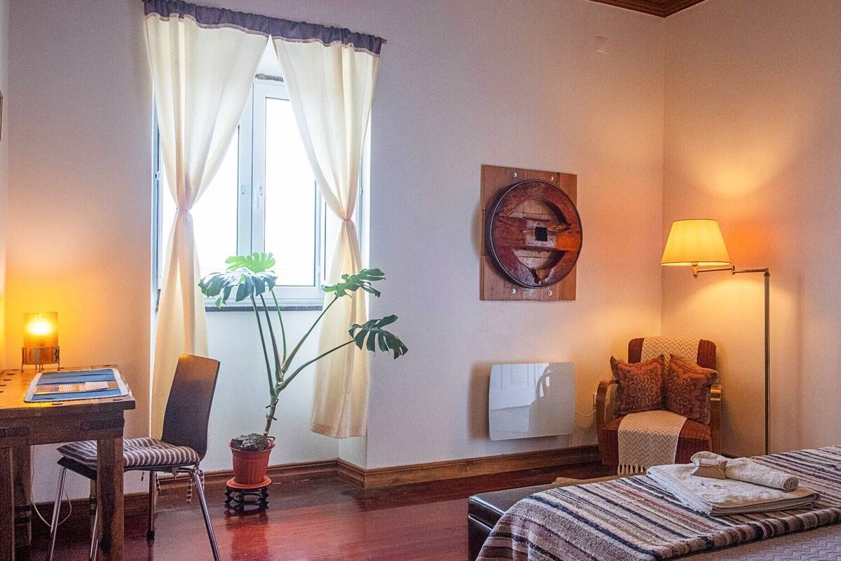 Een gezellige hotelkamer met een bed, een bureau met lamp, een stoel, een potplant en een wandlamp.
