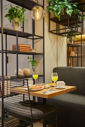 Un'accogliente zona pranzo con un tavolo in legno, sedie in metallo nero e un divano grigio. Si notano libri, un vaso con una pianta e una lampada a sospensione.