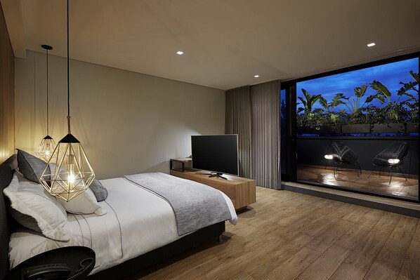 A modern bedroom with a large bed, bedside lamps, a TV, and a view of plants through a large window.