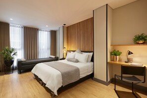 A modern hotel room with a large bed, a desk with a chair, a potted plant, and a window with curtains.