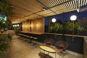 An outdoor bar area with wooden slatted ceiling, bar stools, and round tables.