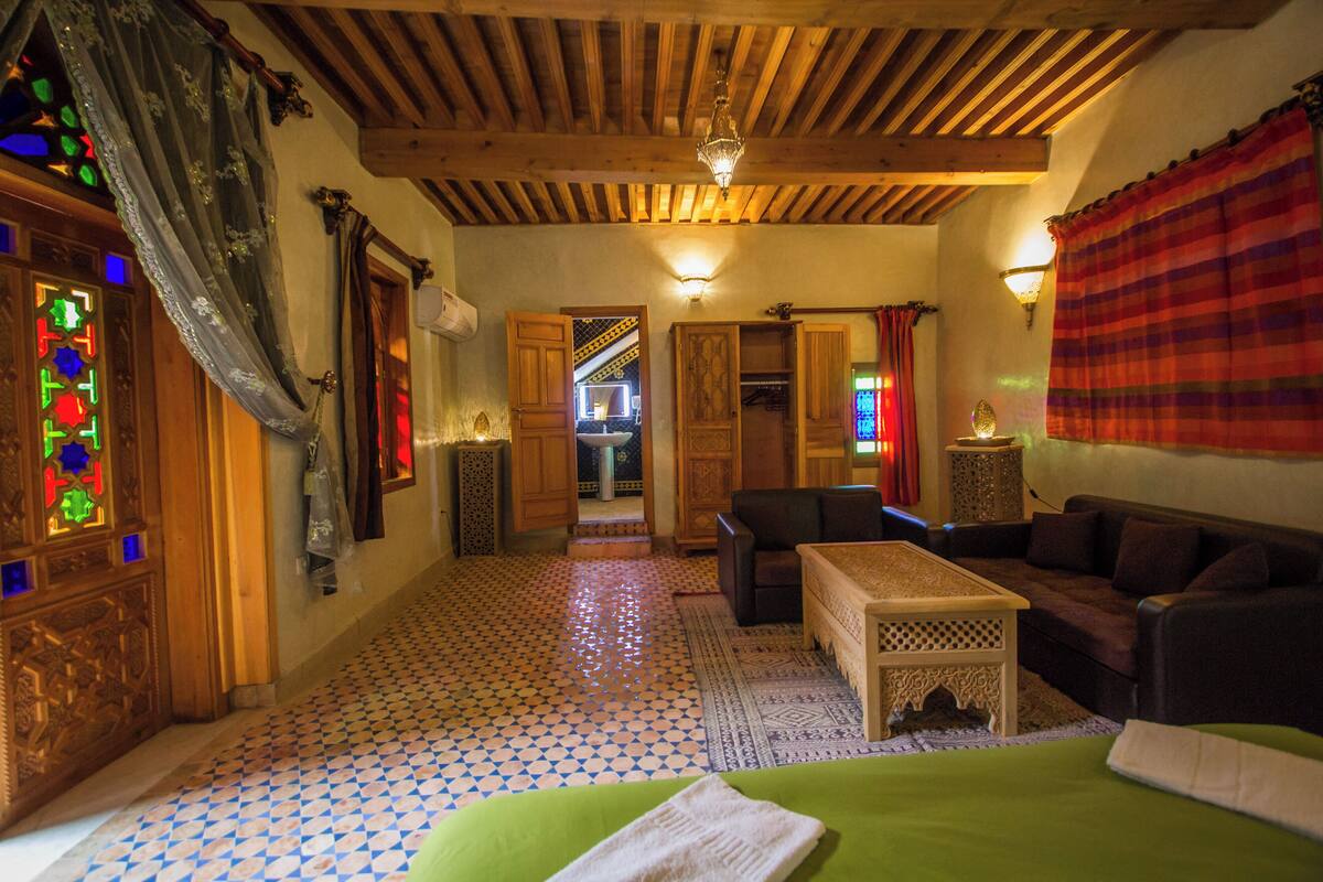 Um quarto acolhedor com piso de azulejos, vigas de madeira no teto, cama verde, mesa de madeira e um sofá.