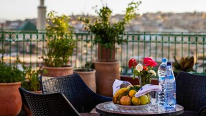 A table with a fruit bowl, flowers, and water bottles on a balcony with potted plants and a view of a cityscape. - Riad Al Fassia Palace (Fes)
