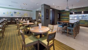 Une salle à manger d’hôtel avec plusieurs tables et chaises, un espace cuisine et un mur où est affiché le menu.