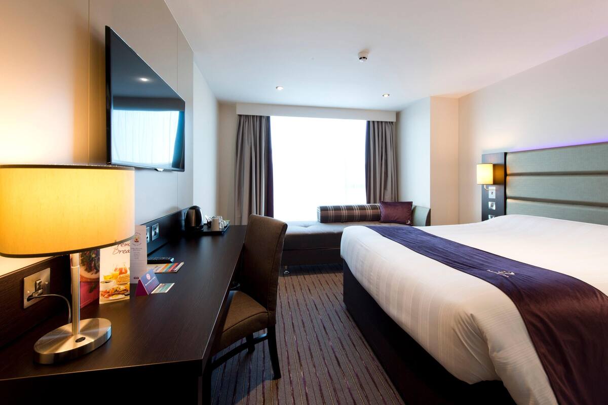 A hotel room with a bed, a desk with a lamp, a TV, and a window with curtains.