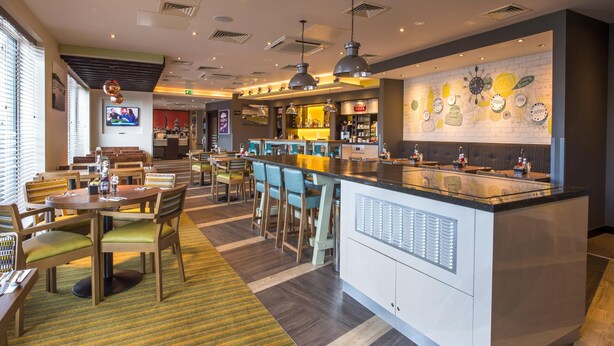 A modern restaurant with a bar area, seating arrangements, and a television screen. - Premier Inn Bude (Bude)
