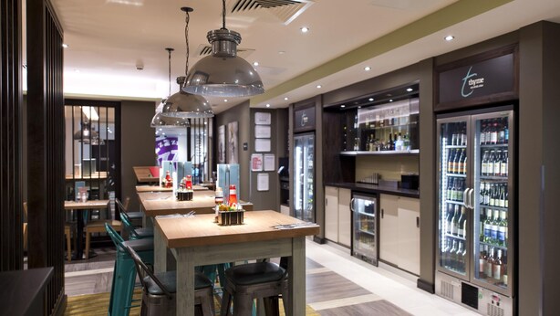 A restaurant with a long counter, bar stools, hanging pendant lights, and a refrigerator stocked with beverages. - Premier Inn London Southgate (London)