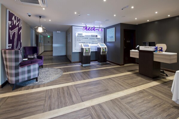 Uma área de recepção de hotel com balcão de check-in, um espaço de estar com poltrona xadrez e um pufe roxo, e uma parede com palavras motivacionais.