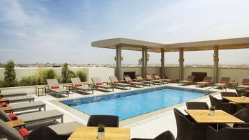 A rooftop pool area with lounge chairs and tables, a clear view of the cityscape, and a covered seating area.