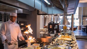 A chef performing a flambé in a modern kitchen with a buffet line of dishes.