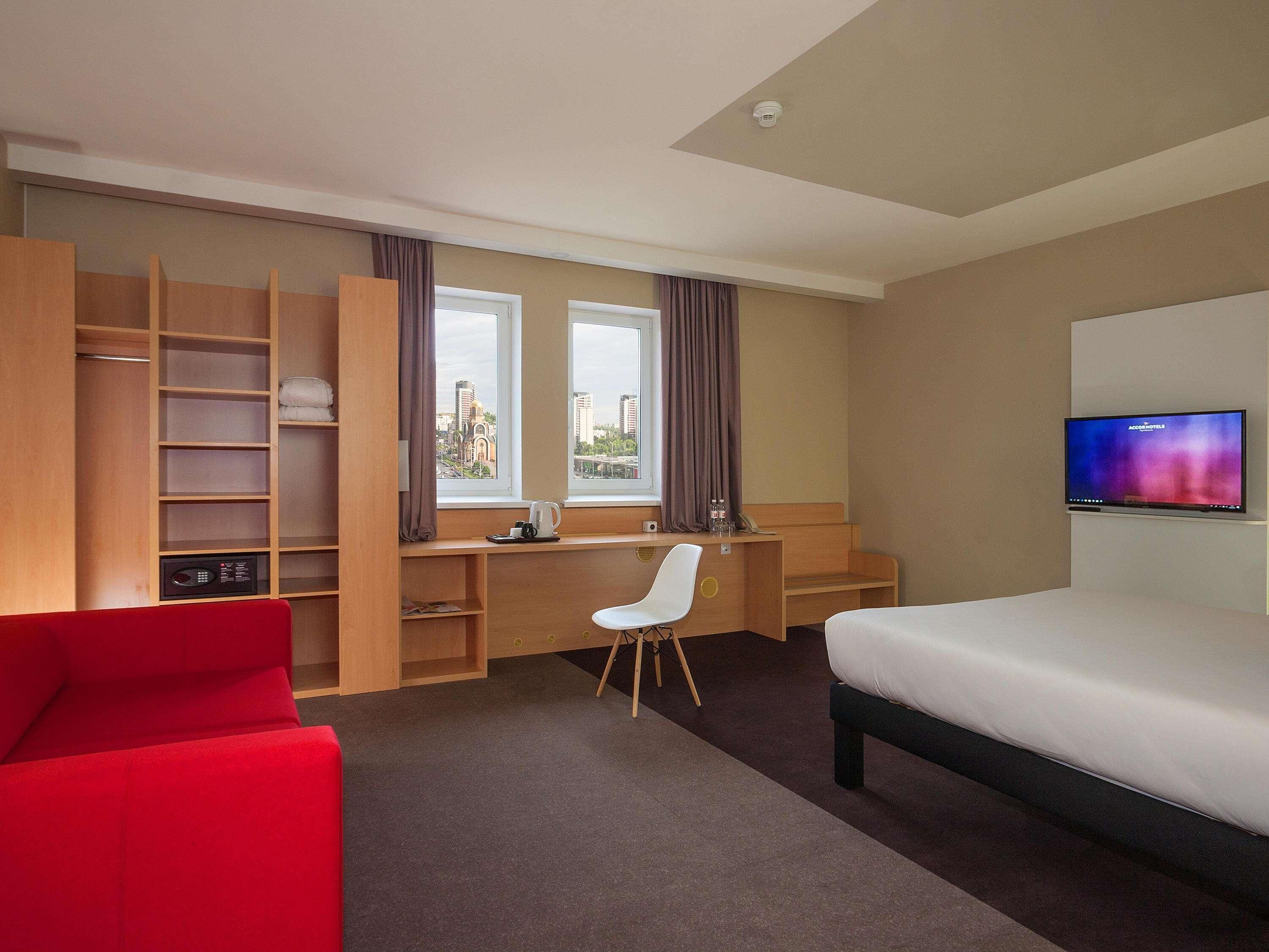 A hotel room with a bed, a desk with a chair, a TV, a red sofa, and a view of buildings through the window.