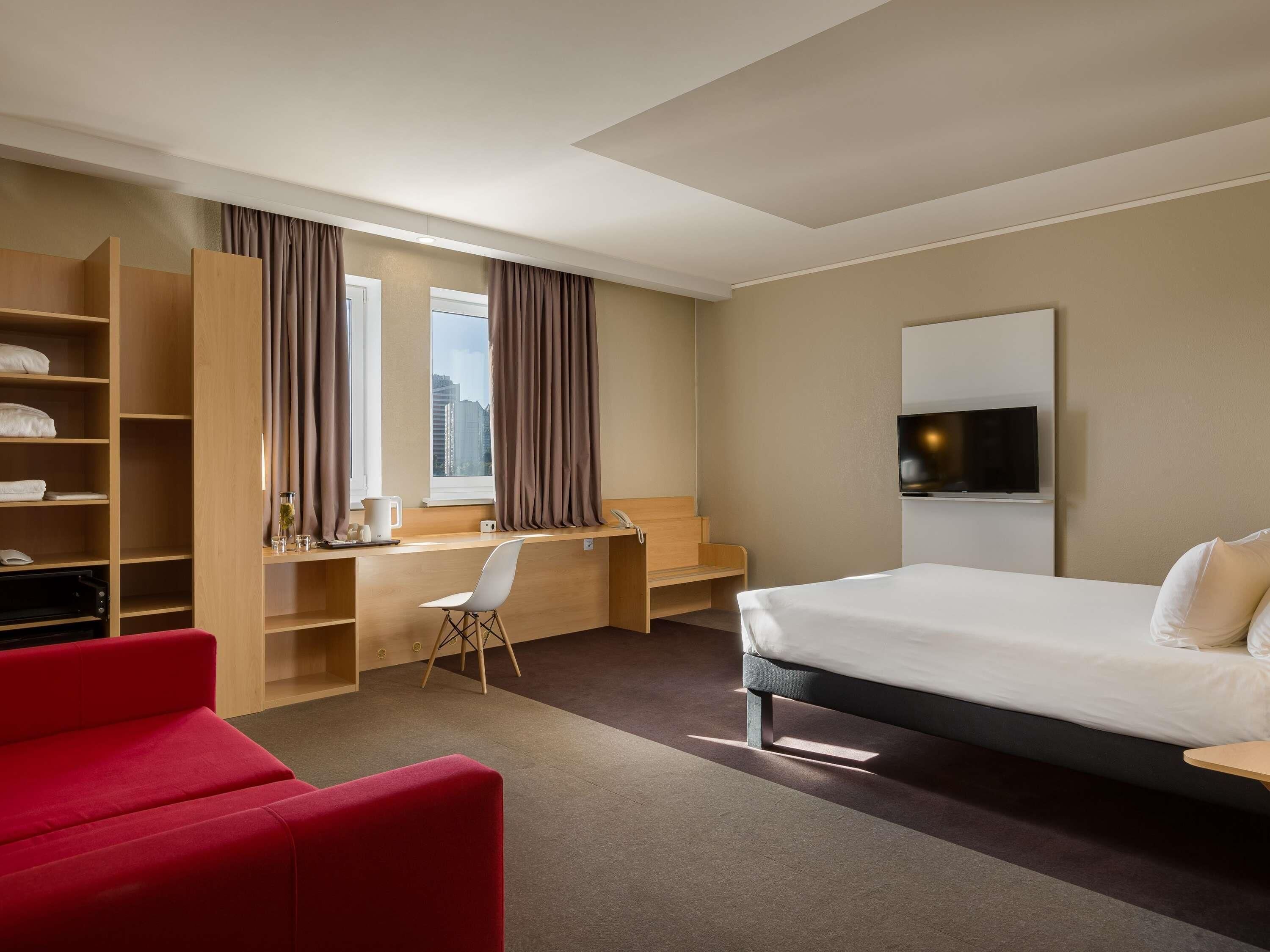 A hotel room with a bed, a desk, a chair, a TV, and a red sofa.