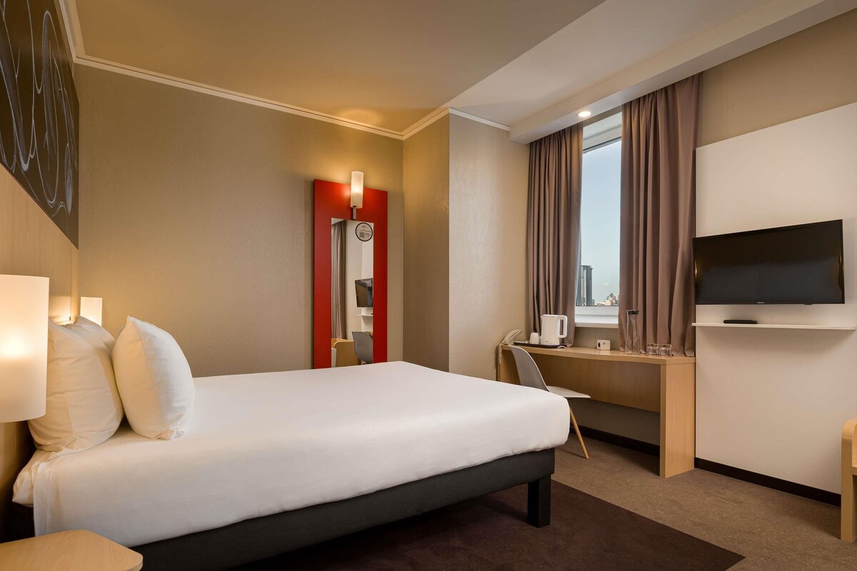 A hotel room with a bed, desk, chair, TV, and a view of the city.