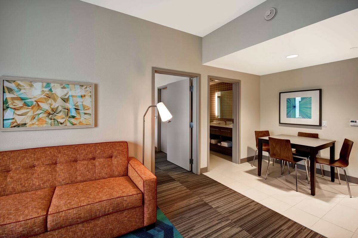 A hotel room with a sofa, a dining table with chairs, a painting on the wall, and a hallway leading to a bathroom.