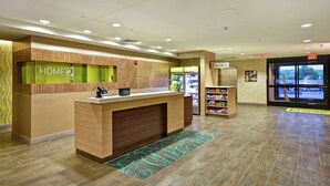 A hotel reception area with a reception desk, a digital display, and a view of the outside. - Home2 Suites by Hilton Mechanicsburg (Mechanicsburg)