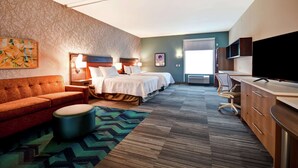 A hotel room with a sofa, two beds, a TV, a desk, and a patterned carpet. - Home2 Suites by Hilton Mechanicsburg (Mechanicsburg)
