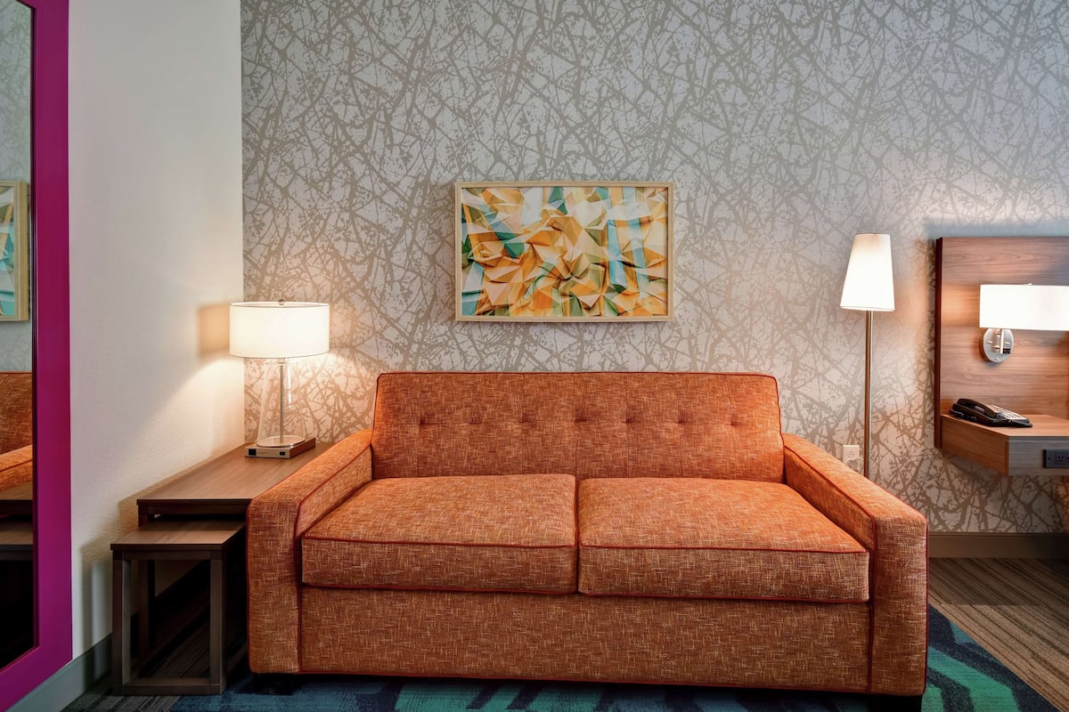 A modern hotel room with a sofa, a side table with a lamp, a wall-mounted artwork, and a wooden nightstand with a lamp.